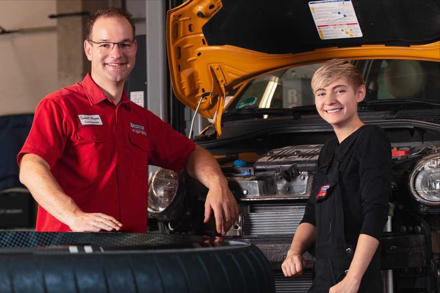 Ein junger Mann steht lächelnd an der offenen Motorhaube eines Autos und blickt in die Kamera. Neben ihm steht eine junge Auszubildende die ebenfalls freundlich in die Kamera blickt.